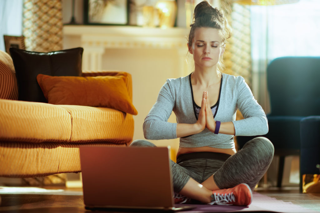 relaxed fit sports woman in fitness clothes in the modern house meditating using online yoga training program in laptop.