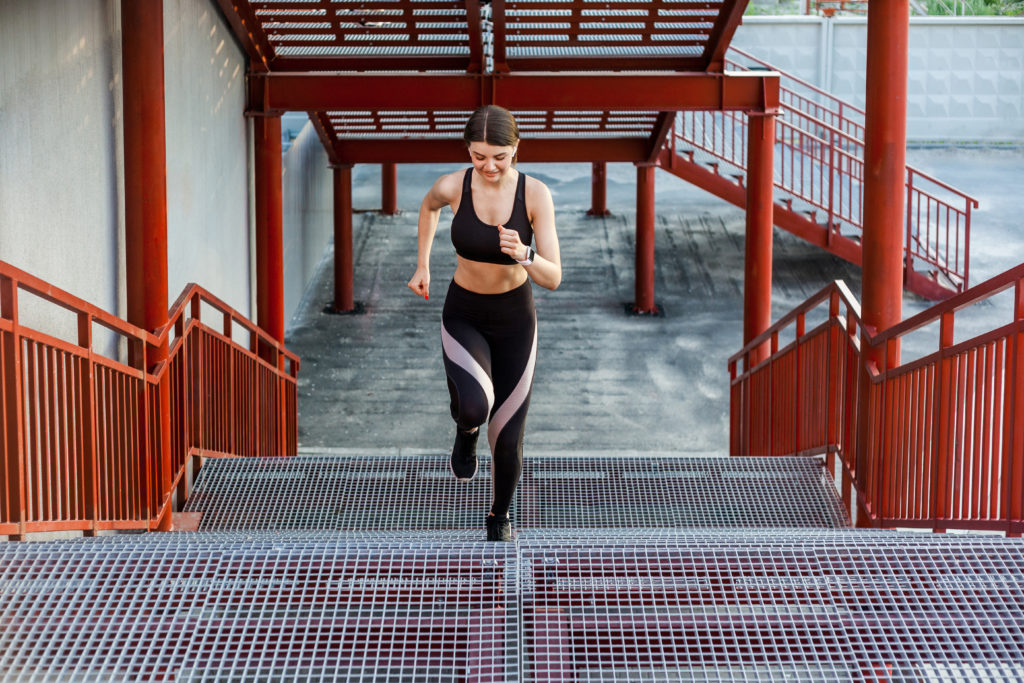 Straight view of young athletic slim beautiful woman running up stairs doing cardio interval training in black fashionable sportwear in the street on summer. Outdoor, sport and healthy concept
