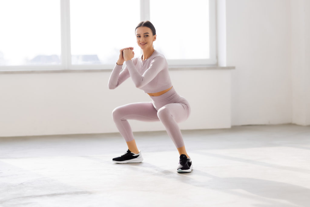 Young woman in sportswear doing squat while standing in front of window at gym