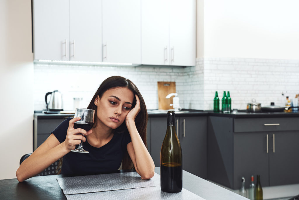 Dark-haired, sad and wasted alcoholic woman sitting at home, in the kitchen, leaning over the table, holding a glass and looking at bottle of red wine, completely drunk, looking depressed, lonely and suffering hangover in alcoholism and alcohol abuse