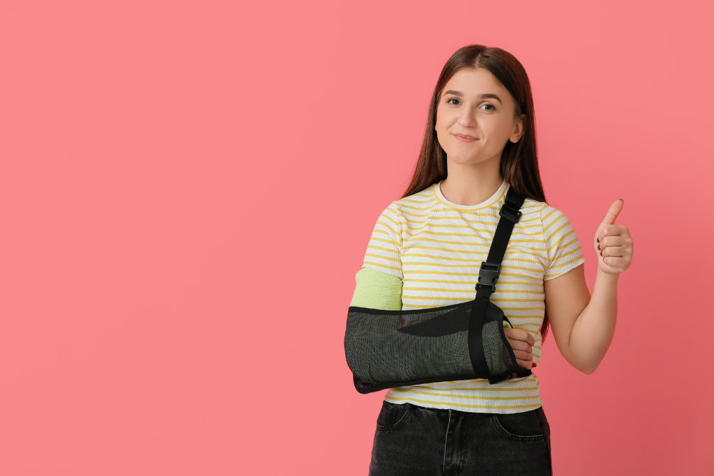 Young woman with broken arm showing thumb-up on color background　肩を吊る