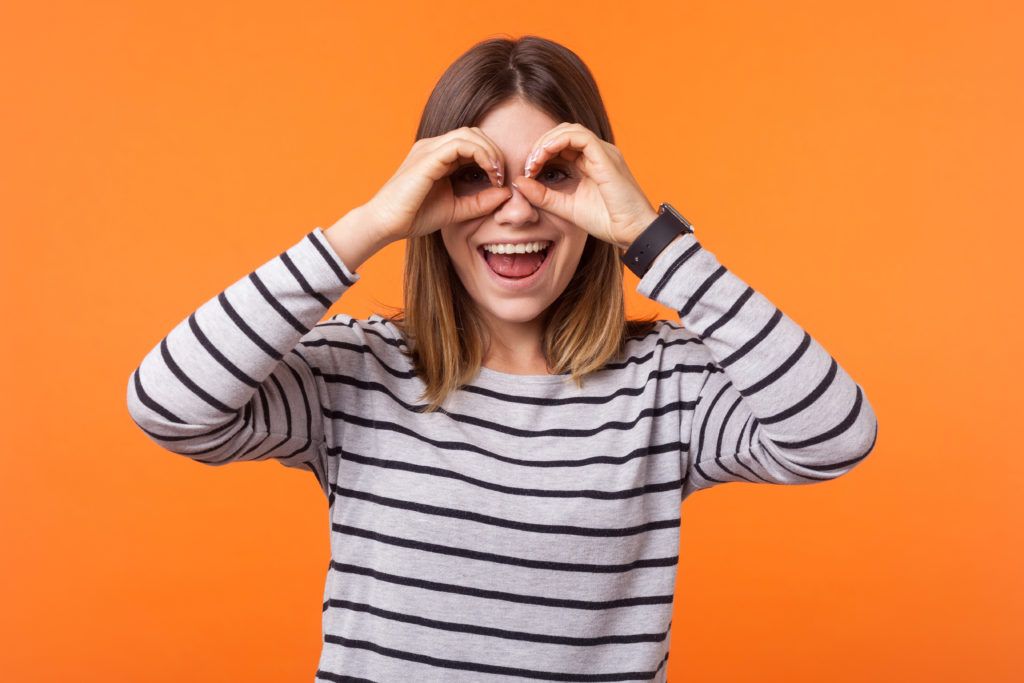 Portrait of curious positive woman with brown hair in long sleeve striped shirt standing, holding hands near eyes like spectacles, binocular gesture. indoor studio shot isolated on orange background