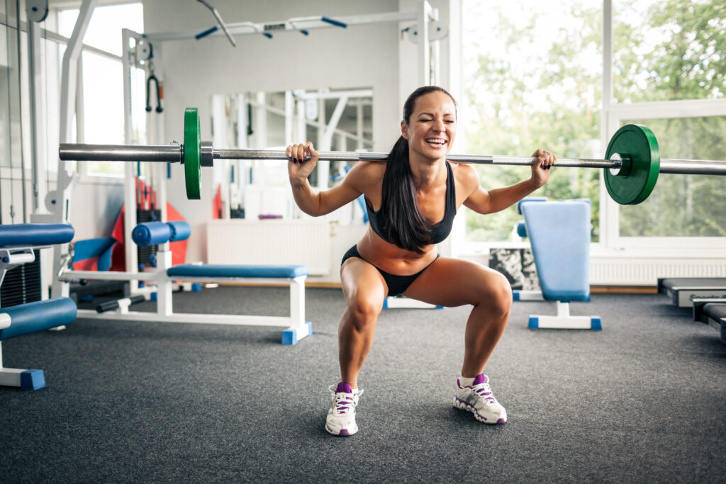 Front view of charming sporty lady doing exercise with barbell in gym. Cheerful athletic girl having strength workout. Concept of sport, crossfit and training.